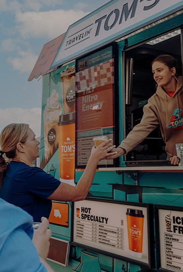 Travelin' Tom's Church Events A woman at a community event is handed a drink from a franchisee working at the window of a Travelin’ Tom’s Coffee Truck