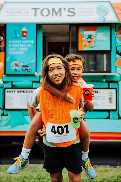 A girl gives a boy a piggyback ride while they pose with iced specialty drinks in front of a Travelin' Tom's Coffee Truck after a sports event