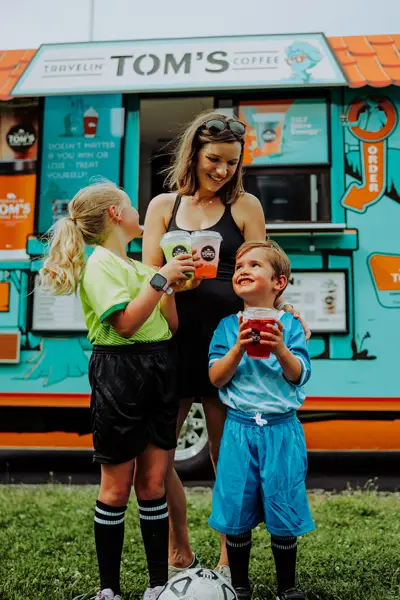 A mom and two children stand in front of a Travelin' Tom's Coffee Truck while enjoying iced beverages after a soccer game