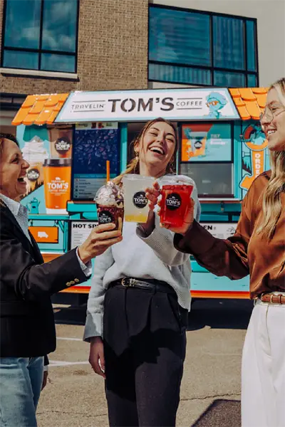 A group of people at an event smile and toast with hot and iced beverages while standing in front of a Travelin' Tom's Coffee Truck
