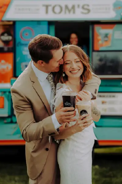 A bride and groom embracing while enjoying iced beverages from a Travelin' Tom's Coffee truck during their wedding celebration