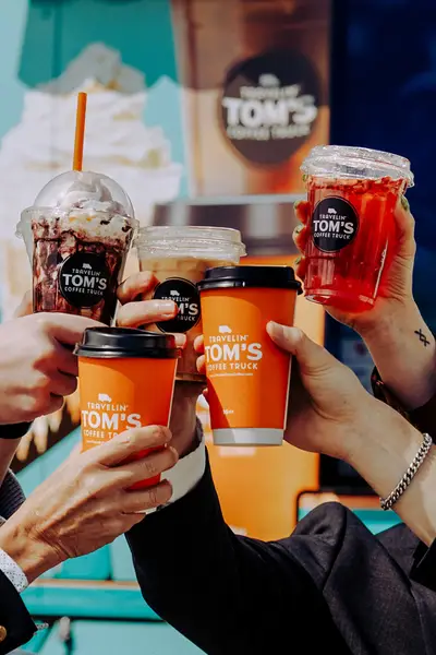 Close-up of five people's hands raising iced, frozen, and hot specialty beverages from a Travelin' Tom's Coffee Truck in a celebratory toast