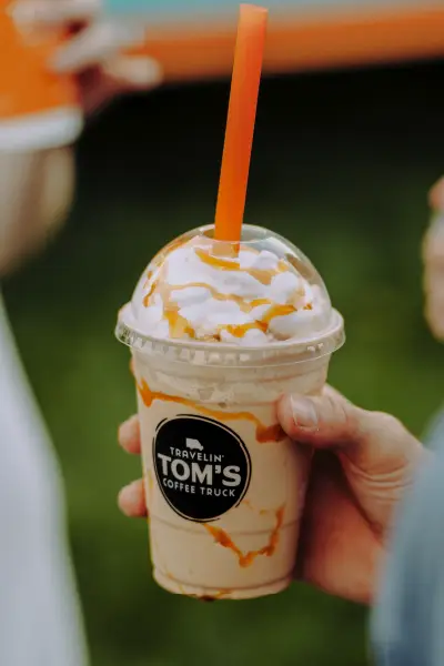 Closeup of a hand holding a Travelin' Tom's caramel frappe topped with whipped cream and caramel drizzle